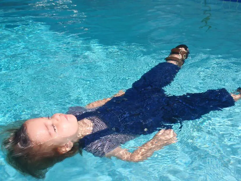 A toddler in long sleeves and trousers floating on her back in a pool